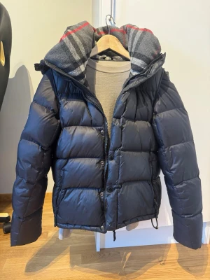 Svart pufferjacka från Burberry - Snygg svart pufferjacka från Burberry med patch på ärmen där det står 'Burberry London England'. Jackan har dragkedja och knappar, samt en avtagbar huva med rutigt foder i grått, rött och vitt. Perfekt för kalla vinterdagar och riktigt trendig look.