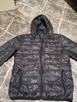 Svart pufferjacka från EA7 Emporio Armani - Snygg svart pufferjacka från EA7 Emporio Armani med huva och dragkedja framtill. Jackan har två sidofickor med dragkedja och diskret logga på bröstet. Perfekt för kalla dagar och ger en sportig vibe.