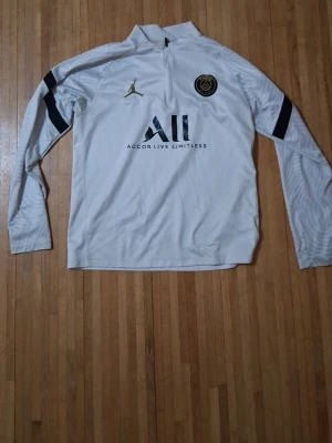 Vit PSG träningströja från Jordan - Säljer en vit långärmad träningströja från PSG och Jordan med svart logga och guldfärgad Jumpman på bröstet. Tröjan har halv dragkedja, svarta detaljer på ärmarna och texten 'Accor Live Limitless' framtill. Perfekt för fotbollsträning eller chill.
