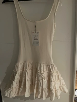 Vit volangklänning med smala axelband - Supersöt vit kortklänning med smala axelband och flera volanger nertill. Klänningen har en enkel, figurnära överdel och en fluffig kjol med volangdetaljer. Perfekt för sommarens alla tillfällen.