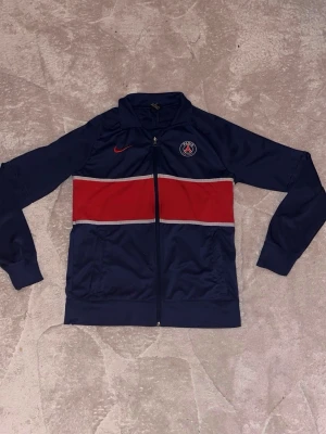 PSG träningsjacka från Nike - Mörkblå träningsjacka från Nike med PSG-logga på bröstet och röd Nike-swoosh. Jackan har en bred röd rand över bröstet med vita detaljer, hel dragkedja och ståkrage. Perfekt för fotbollsträning eller som sportig streetwear.