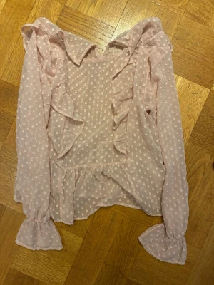 Ljusrosa prickig blus med volanger - Söt ljusrosa blus med vita prickar och volangdetaljer framtill. Blusen har långa ärmar  och volang 