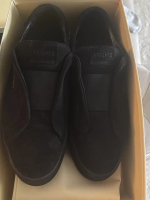 Svarta sneakers från Axel Arigato - Stilrena svarta sneakers från Axel Arigato med minimalistisk design. Skorna har en bred elastisk rem över vristen och är tillverkade i mocka. Perfekta för dig som gillar en clean och modern look.