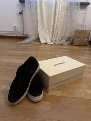 Svarta sneakers från Axel Arigato - Säljer ett par svarta sneakers från Axel Arigato i mocka med vit platt sula. Skorna har minimalistisk design utan snörning och elastiska paneler för enkel påtagning. Märket syns diskret på sidan och tungan. Perfekta för dig som gillar stilrena och bekväma skor. Lite slitage i hälen på vänstra skon men inget man märker av