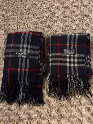 2st burberry halsdukar - Två klassiska halsdukar från Burberry i mörkblått med rött och vitt rutmönster. Båda har fransar i ändarna och märkets logotyp invävd. Perfekt accessoar för att lyfta din outfit med en tidlös brittisk stil. Materialet är mjukt och känns lyxigt. Billigare än att köpa enskilda!