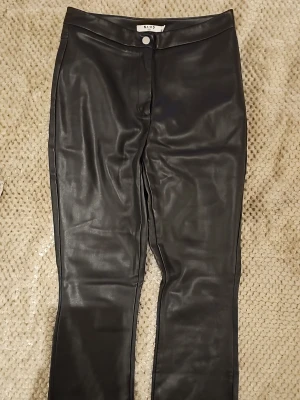 Svarta fuskskinnjeans från NA-KD - Snygga svarta jeans i fuskskinn från NA-KD. De har en rak passform, hög midja och klassisk knappstängning framtill. Perfekta för dig som vill ha en edgy look med skinnkänsla. Materialet är glansigt och mjukt, vilket ger en modern vibe.