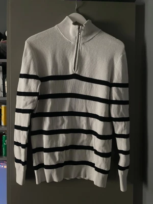 Vit stickad tröja med svarta ränder - Vit stickad tröja med svarta horisontella ränder och halv dragkedja vid halsen. Tröjan har ribbstickad struktur, hög krage och långa ärmar. Perfekt för dig som gillar klassisk och stilren design.