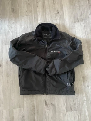 Svart vindjacka från Sail Racing - Svart vindjacka från Sail Racing med dragkedja framtill och hög krage med mjukt foder. Jackan har logga broderad på bröstet och justerbara ärmslut med kardborre. Materialet är slitstarkt och vattenavvisande, perfekt för blåsiga dagar.