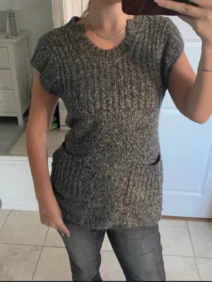 Gråmelerad stickad väst med fickor - Säljer en gråmelerad stickad väst med korta ärmar och två praktiska fickor framtill. Västen har ribbstickad struktur och rund halsringning. Perfekt att slänga över en långärmad tröja för extra lager och stil.