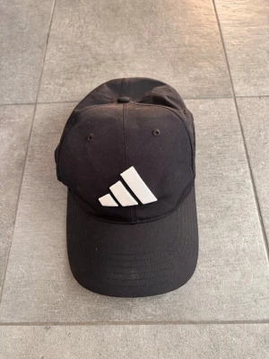 Svart keps från Adidas - Svart keps från Adidas med klassisk böjd skärm och justerbar passform. Kepsen har en stor vit Adidas-logga framtill. Perfekt accessoar för en sportig och avslappnad stil.