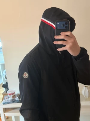Svart Moncler vindjacka med huva - Snygg svart vindjacka med huva och dragkedja. Jackan har Moncler-logga på vänster ärm och en vit-röd rand på huvan. Perfekt för dig som vill ha en stilren och sportig look. Materialet är tunt och vindtätt, passar bra till vår och höst.
