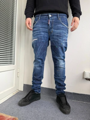 DSQUARED2 - Säljer ett par blå jeans DS2 med avslappnad passform och färgstänk. Baksidan har en stor röd bladpatch på ena bakfickan och coola slitningar framtill. Jeansen har klassiska fem fickor och knappgylf. Perfekta för dig som gillar streetwear och unika detaljer. Han på bild är 175cm och väger 74kg
