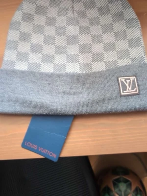 Grå Louis Vuitton mössa med rutmönster - Snygg grå mössa från Louis Vuitton med klassiskt rutmönster och uppvikt kant. LV-logga framtill och diskret etikett på insidan. Perfekt accessoar för att lyfta din stil under kyliga dagar.