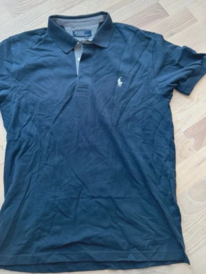 Mörkblå piké från Ralph Lauren - Klassisk mörkblå pikétröja från Ralph Lauren med vit broderad logga på bröstet. Tröjan har krage, knappar vid halsen och rutigt tyg på insidan av knappslån. Perfekt till en avslappnad men ändå stilren look.