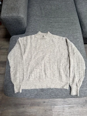 Beige ribbstickad tröja med hög krage - Säljer en beige ribbstickad tröja med högre krage och långa ärmar. Tröjan har breda muddar vid ärmslut och nederkant, och är gjord i ett mjukt, tjockt material. Den är från Grunt i storlek M men passar även S. Den är inte andvänd mer än 2 gånger.