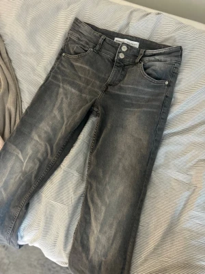 Grå jeans från Bershka - Säljer ett par grå jeans från Bershka i storlek 38. Jeansen har en klassisk femficksmodell, lite utsvängda ben och snygga slitningar för en cool look. Tillverkade i bomullsblandning och stängs med dragkedja och dubbla knappar.