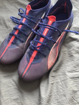 Blå och röda Puma fotbollsskor - Säljer ett par snygga fotbollsskor från Puma i blått med röda detaljer och vita inslag. Skorna har snörning, låg profil och är tillverkade i ett lätt syntetmaterial för bra bollkontroll. Perfekta för dig som vill sticka ut på planen!