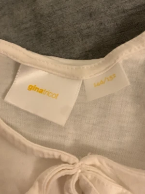 Vit blus med puffärm från Gina Tricot - Söt vit blus från Gina Tricot med puffiga korta ärmar och volang nedtill. Blusen har en rund halsringning och knytning framtill som ger en extra detalj. Perfekt för dig som gillar en feminin och trendig stil.