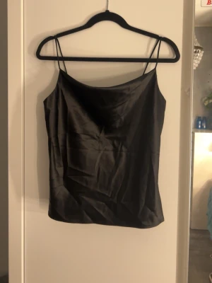 Svart satin linnetopp med smala band - Stilren svart linnetopp i satin med tunna axelband och rak passform. Perfekt att styla med jeans eller kjol för en trendig look. Materialet ger ett snyggt, glansigt intryck och toppen är ärmlös för en luftig känsla.