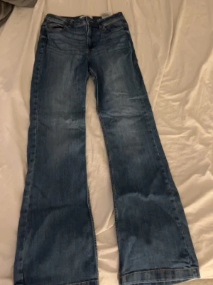 Blå bootcut jeans med slitningar - Säljer ett par blå bootcut jeans med lätt slitna detaljer och klassisk femficksdesign. Jeansen har normal midja och är tillverkade i denim med en snygg tvätt. Perfekta för dig som gillar en avslappnad men trendig look.