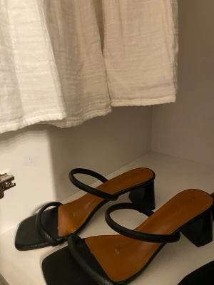 Svarta sandaler med klack och remmar - svarta låga klackar med fyrkantig tå, två tunna band över foten. Klackarna har en låg, stabil klack och innersula i brunt. Perfekta för dig som letar efter ett par med låg klack och vill ha något som matchar till allt. Har kommit till användning 1 gång pgr av att dem är lite för stora. 