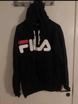 Svart hoodie från FILA - Svart hoodie från FILA med stor vit och röd logga framtill. Klassisk modell med huva och vita dragsnören. Perfekt för en avslappnad och sportig stil. Mjuk bomullskänsla och rymlig passform.