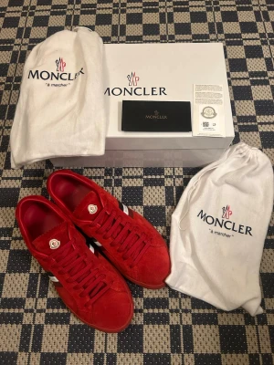 Röda Moncler sneakers i mocka - Snygga röda sneakers från Moncler i mjuk mocka med vita och svarta detaljer på sidan samt Moncler-logga på plösen. Skorna har klassisk låg profil och matchande röda skosnören. Kommer med originalkartong och dustbags.