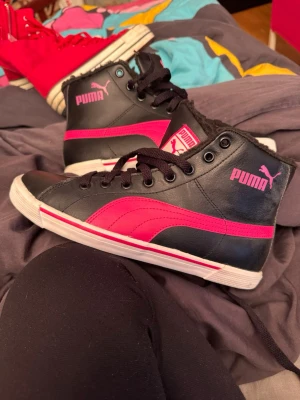 Svarta och rosa Puma high top sneakers - Snygga high top sneakers från Puma i svart och rosa. Skorna har klassisk Puma-logga på sidan och tungan, svarta skosnören och vit sula med rosa detaljer. Tillverkade i syntetmaterial och läderlook för en cool streetstil.är mera som en 36 i storlek 