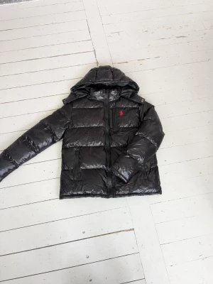Svart pufferjacka från Polo Ralph Lauren - Snygg svart dunjacka från Polo Ralph Lauren med blank finish och röd logga på bröstet. Jackan har huva, dragkedja framtill och två stora fickor med dragkedja. Perfekt för kalla vinterdagar och riktigt skön att bära.1.1 kopi. Storlek S men är som xs. Helt nytt skick och har knappast blivit använd. ENDAST KÖP VIA POSTNORD