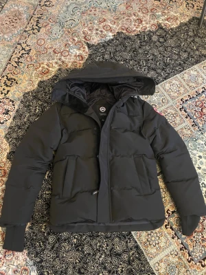 Canada Goose jacka - Snygg svart dunjacka från Canada Goose med huva och två stora framfickor. Jackan har en klassisk puffer-modell och patch med logga på ärmen. Perfekt för kalla dagar och riktigt varm tack vare dunfyllningen.