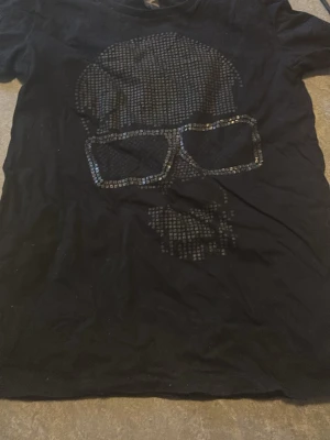 Svart t-shirt med döskalletryck - Cool svart t-shirt med ett stort döskalletryck framtill i glansiga svarta rutor. T-shirten har klassisk passform och korta ärmar. Perfekt för dig som gillar edgy och unik stil.