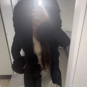 Svart pälsjacka med stor krage - Snygg svart pälsjacka med extra stor fluffig krage och muddar i fuskpäls. Jackan har en rak passform och stängs framtill. Perfekt för kalla dagar när du vill ha en lyxig look.