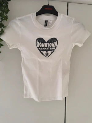 Vit t-shirt Downtown Manhattan H&M, XS - Vit t-shirt från H&M med svart tryck i form av ett hjärta och texten 'Downtown Manhattan' på bröstet. Klassisk rund halsringning och korta ärmar. Perfekt för en avslappnad streetstyle-look.