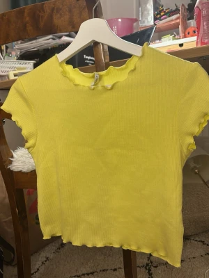 Gul ribbad topp med vågig kant storlek S, men funkar om man är xxs och xs - Snygg gul ribbad topp med korta ärmar och vågiga kanter vid hals, ärmslut och nederkant. Perfekt för dig som gillar färgglada och trendiga plagg. Materialet är stretchigt och mjukt för en bekväm passform.