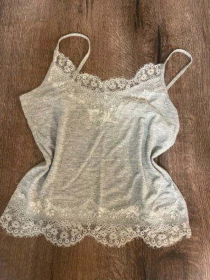 Grått linne med spetsdetaljer - Snyggt grått linne med tunna axelband och spetsdetaljer i vitt. Mjukt material och går att ha till både vardags och som pyjamas. Perfekt att bära under en skjorta eller som den är. Storlek S men passar även en XS. Använt flertal gånger.