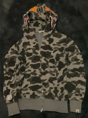 BAPE camo zip hoodie med hajmotiv - Säljer en grå camo zip hoodie från BAPE med ikoniskt hajmotiv på huvan och färgglada detaljer. Hoodien har dragkedja hela vägen upp, ribbade muddar och fickor fram. Materialet är mjuk bomullsmix och passformen är relaxed. Perfekt för dig som gillar streetwear och vill sticka ut.