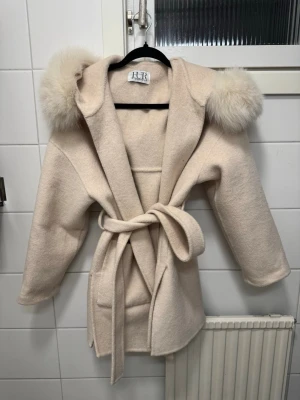Beige kappa med fuskpäls från FUR - Stilren beige kappa från FUR  med stor huva kantad av fluffig fuskpäls. Kappan har knytskärp i midjan och en mjuk, ullig känsla. Perfekt för dig som vill ha en trendig och mysig look under kyliga dagar.