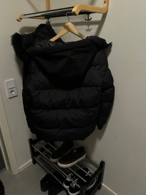 Svart moncler  pufferjacka med luva ny - Säljer en svart pufferjacka med stor luva och fuskpälsdetalj. Jackan har en quiltad design, dragkedja och tryckknappar framtill samt Moncler-logga på ärmen. Perfekt för kalla vinterdagar och riktigt snygg till streetwear-stilen.
