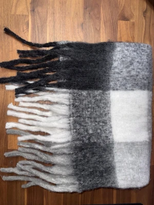 Rutig halsduk i svart, grått och vitt - Mjuk och fluffig halsduk med rutigt mönster i svart, grått och vitt. Halsduken har långa fransar i båda ändar och är perfekt för att hålla värmen under kalla dagar. Materialet känns ulligt och ger en mysig känsla. Halsduken liknar ”Acne Studios halsdukarna och är oanvänd (ny).