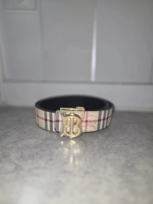 Rutig Burberry-inspirerad bälte - Snyggt bälte med klassiskt rutigt mönster i beige, svart och rött samt ett stort guldfärgat B-spänne framtill. Perfekt accessoar för att lyfta vilken outfit som helst med en touch av lyxig känsla.