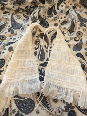 Vit omlott halterneck linnetopp - Supersöt vit omlottopp med halterneck och knyt i ryggen. Toppen har volangkant nertill och är gjord i ett tunt, luftigt material som passar perfekt till sommaren. Perfekt för att skapa en trendig och avslappnad look.