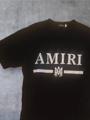 AMIRI t shirt - Svart t-shirt från AMIRI med stor vit logga och grafisk detalj framtill. Klassisk rund halsringning och korta ärmar. Perfekt för dig som gillar streetwear och vill sticka ut med en exklusiv look.