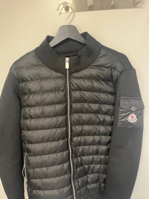 Svart cardiganjacka från Moncler - Snygg svart cardiganjacka från Moncler med quiltad framsida och mjuka stickade ärmar. Jackan har dragkedja framtill, ribbad krage och en ficka med Moncler-logga på ärmen. Perfekt för lager-på-lager och streetstyle.