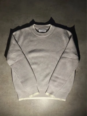 Ljusgrå stickad tröja från maison margela  - Ljusgrå stickad tröja från masion margiela med rund halsringning och ribbade muddar i vitt vid ärmslut och nederkant. Tröjan har en clean och minimalistisk look, perfekt för lager på lager och enkel att matcha med olika outfits. Storlek L och använd men inga defekter. Kom gärna med bud och det är bara att skriva över funderingar!