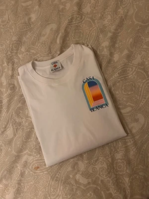 Vit Casa Blanca t-shirt med tryck - Säljer en fräsch vit t-shirt från Casa Blanca med ett färgstarkt grafiskt tryck på bröstet. T-shirten har klassisk passform och rund hals. Perfekt för dig som gillar stilrena plagg med en twist. Storlek L men passar även M