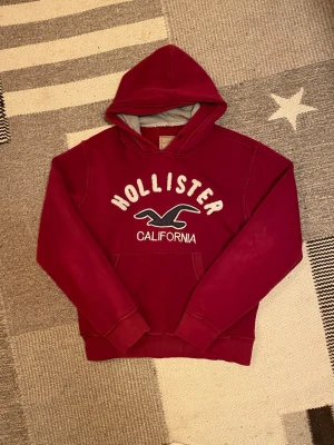 Vinröd hoodie från Hollister - Röd limiterad Hollister hoodie med ”Hollister Californa” logga på bröstet. Tröjan är i gott skick utan fläckar eller större defekter. Hör av dig vid minsta fundering!🙌😁