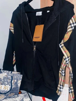 Svart Burberry hoodie med dragkedja - Svart hoodie från Burberry med dragkedja framtill och klassiska beige-rutiga paneler längs ärmarna. Huva med dragsko och två fickor framtill. Tillverkad i mjukt bomullsmaterial för en skön och avslappnad look. Perfekt för dig som vill ha en stilren hoodie med lyxig känsla.