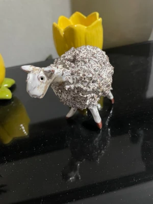 Dekorativ fårfigur i keramik - Söt fårfigur i keramik med lockig, vit och grå päls och detaljerade drag. Perfekt som prydnad till hyllan eller skrivbordet. Fåret har små bruna detaljer på benen och ett uttrycksfullt ansikte.