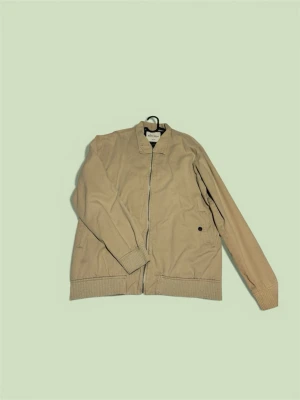 Beige bomberjacka XL/L - Stilren beige bomberjacka från Lager 157 med dragkedja framtill, ribbade muddar och två sidofickor. Jackan har en klassisk bomberkrage och är tillverkad i ett mjukt bomullsmaterial som ger en avslappnad look.