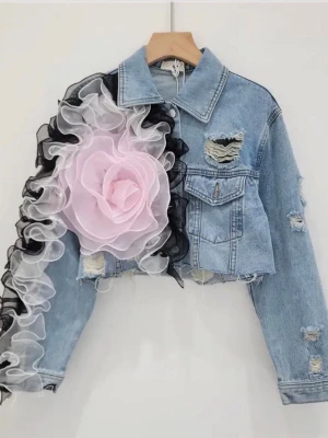 kort jeansjacka med mesh-patchwork och slitna detaljer - Trendig ljusblå jeansjacka med croppad passform och slitna detaljer. Ena ärmen är dekorerad med en stor rosa blomma i organza och svarta samt vita volanger. Jackan har klassisk krage och bröstfickor med lock.